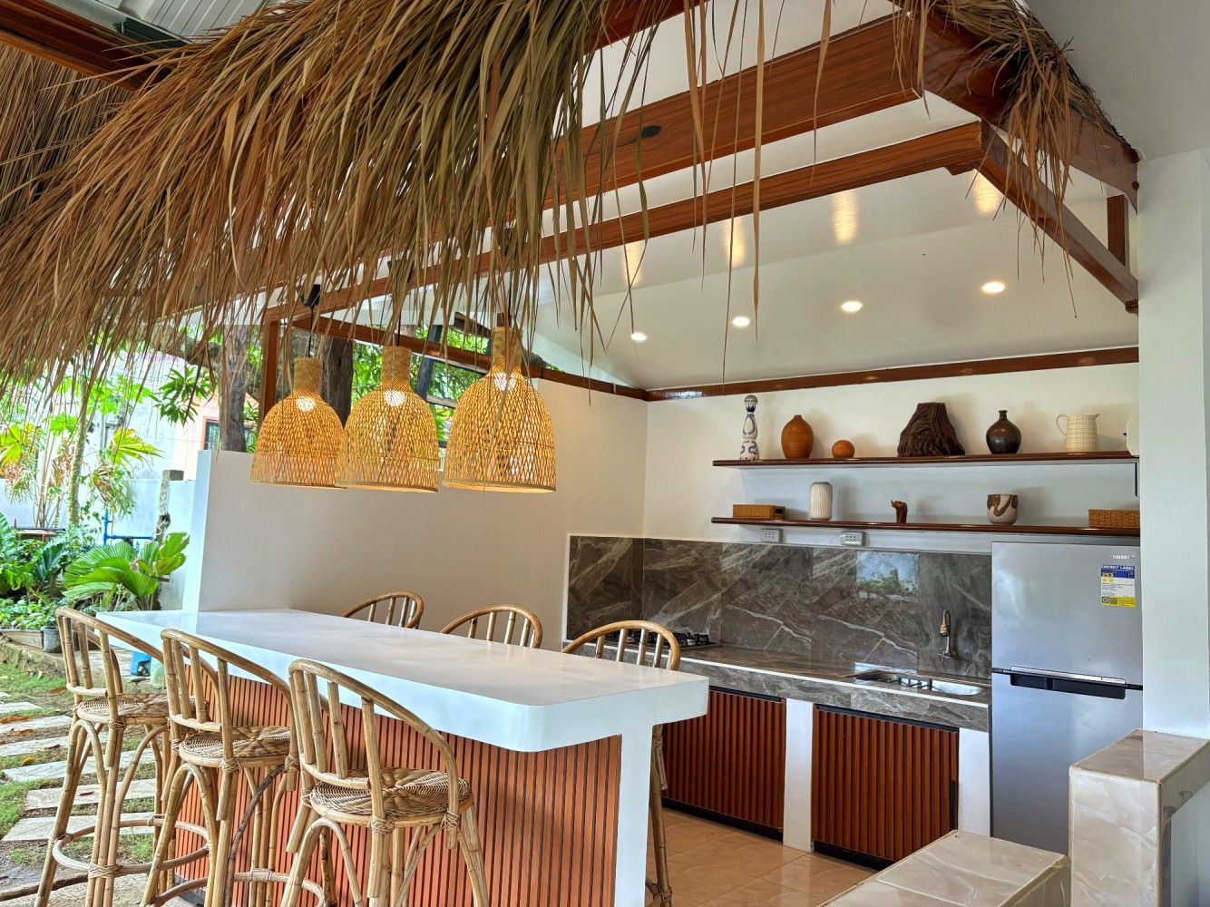 Outdoor kitchen and bar under a thatched roof, with rattan pendant lamps and bar stools
